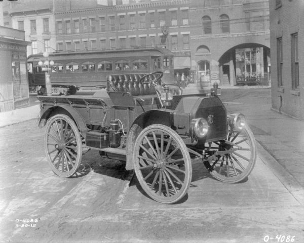 An early IH automobile is parked in a street. The automobile features lamps, possibly gas lamps, as well as the IHC logo on the grill. In the background are storefronts, pedestrians, and a streetcar.