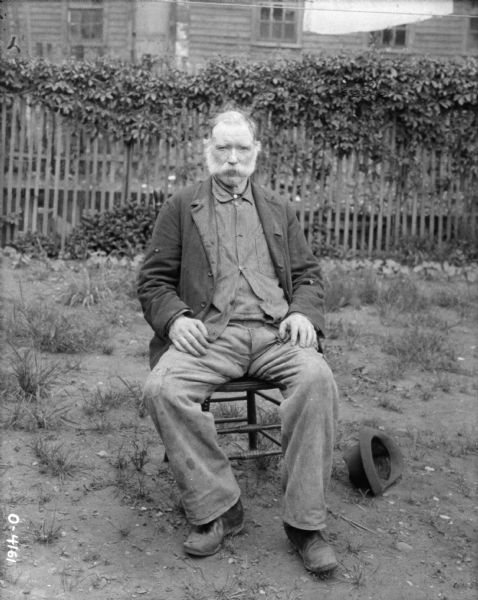 Portrait of a man sitting outdoors in a chair. There is a hat on the ground next to the chair. In the background is a fence and a building.