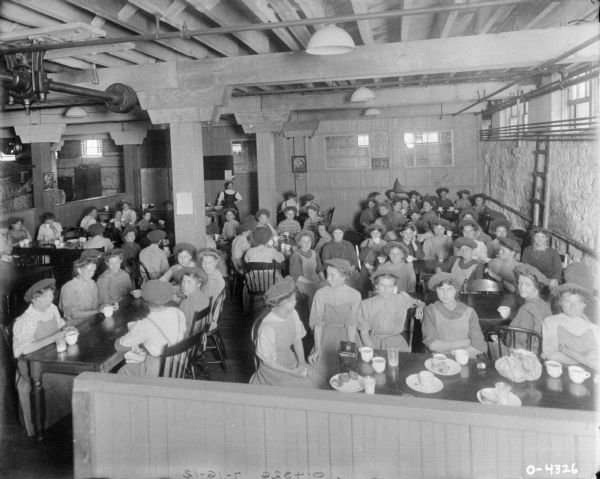 Slightly elevated view of a canteen filled with women dressed in work clothes and hats. The room is equipped with wooden tables, chairs, and white dishes. The room appears to be on a lower level of a factory as windows are along the top of the room. The wall has a few posters of religious figures. In a back corner is a women wearing a dark apron with a cart of food. The women are likely employees of the International Harvester Osbourne Works in Auburn, New York.