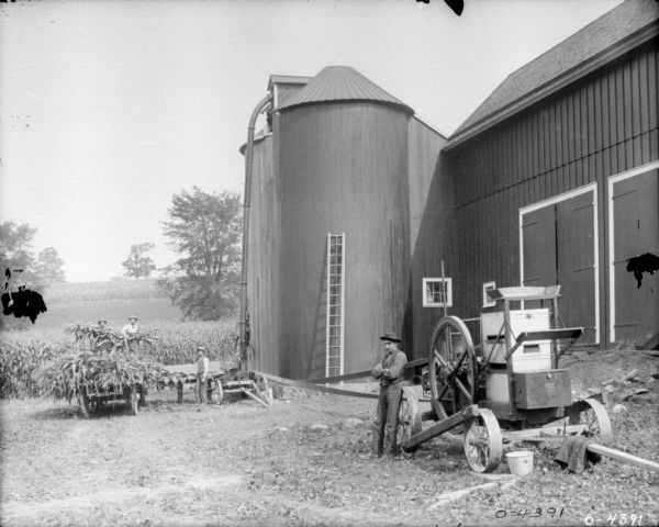A man on the right is standing and leaning against a large cart which contains a vertical stationary engine. Three men on the left are working in the background with an ensilage cutter, harvesting what appears to be corn from a wooden cart, near a large barn and silo. The men are all wearing work clothes and hats. This photograph was probably taken in Auburn, New York.