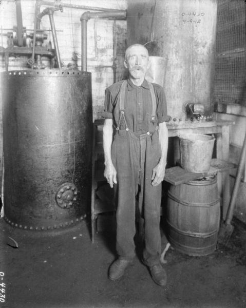 An elderly man is standing and posing inside a factory room. Behind him on the left is a large tank, and on the right is a tank on a table with a barrel underneath the spout.