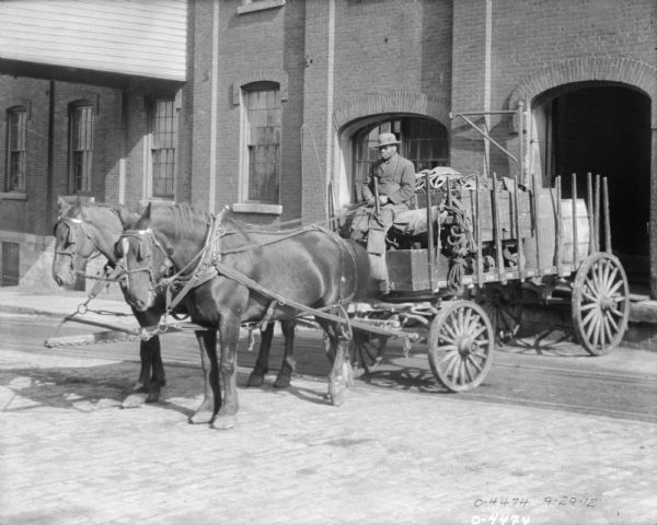 A man is driving a horse-drawn delivery cart, loaded with parts, leaving the plant. Behind the cart is an open arched doorway.