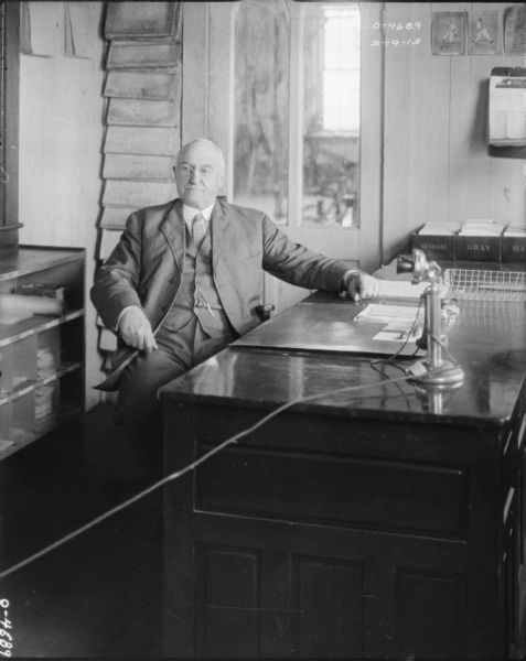 Portrait of a man in a business suit posing behind a desk. A cord in the foreground is attached to a telephone on the desk.