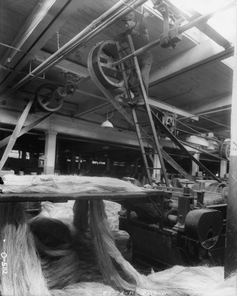 A man is standing on a ladder up near the exposed ceiling of a factory. He is looking at belt driven machinery running twine manufacturing machines.