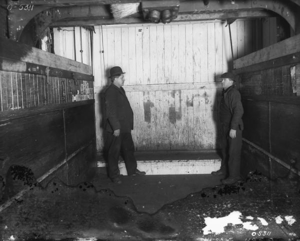 Two men are standing and facing each other at the back of a freight elevator. On the left side of the elevator are tools hanging from nails, and postcards and other printed images are posted nearby. On the right side of the elevator is a sign that reads, in part: "21 Capacity 6000  Lbs."