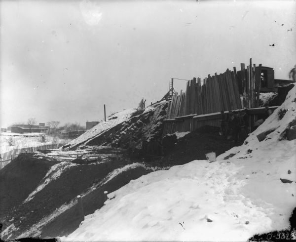 View along side of a hill that is being worked into levels, with snow on the ground. Men, some holding shovels, are standing near the base of a temporary vertical fence which is holding the dirt at the top of the hill in place. The cab of what may be a crane is to the right of the temporary fence. There are fences and buildings in the background on the left.