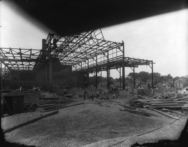 View across pile of gravel towards men standing among the steel structure on a construction site. Lumber is piled on the right, and houses are in the background.