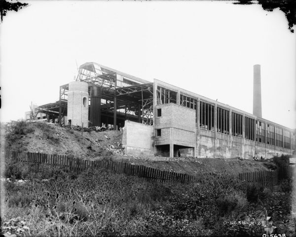 View looking uphill towards a factory building under construction. A brick chimney is near a tall metal tower on the left side of an open steel or iron structure. Men are working along the base of the cement wall. A smokestack is in the background on the right.
