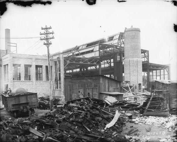 View of construction site of factory building. Metal parts are stacked in the foreground, and men are sitting in a railroad car on the left. A man is at the to of a tall ladder near the brickwork of the new factory building. The rest of the factory building has an exposed steel or iron structure, and on the right men are working on the top of a brick chimney which may be for a forge. A tall metal boiler next to it has a sign that reads: "Manufactured by The Wickes Boiler Company, Saginaw, Michigan, U.S.A."