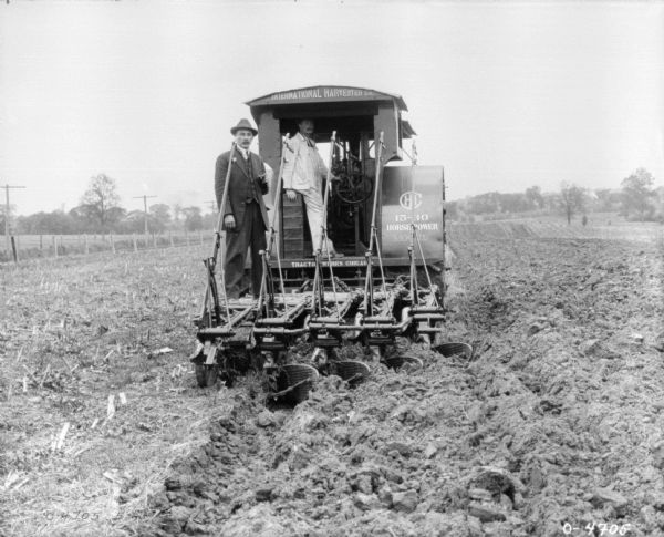 Rear view of two men standing on the back of an IH tractor pulling a plow in a field. The signs on the tractor read: "IHC 15-30 Horse Power Mogul" and "Tractor Works Chicago."