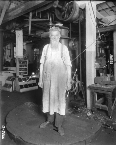Fred Van Patten, wearing a work apron, is standing on a wooden platform in front of a machine.