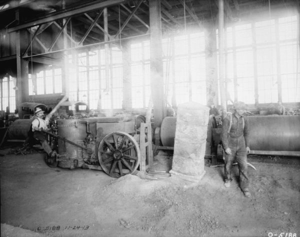 A man on the left is sitting on an early tractor that appears to be equipped with a fork lift, and is moving parts around a factory. Another man is standing on the right. Both men are dressed in work clothes and hats, and have moustaches. In the background is machinery. The men are likely employees of the International Harvester Osbourne Works in Auburn, New York.