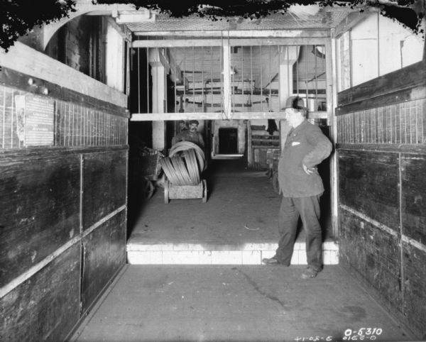 One man wearing a jacket and hat is standing in a freight elevator with  a partially open door, while a second man is pushing a cart of parts toward the elevator. The men are likely employees of the International Harvester Osbourne Works in Auburn, New York.