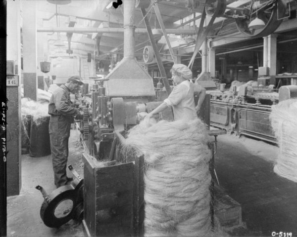A woman dressed in a uniform and puffy hat is standing and operating a large twine machine. To her left is a man dressed in work clothes who appears to be working on the machine. In the background are several other pieces of machinery and electric lights hanging from the rafters. These workers are likely employees of International Harvester Osbourne Works in Auburn, New York.