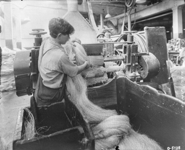 Man operating twine manufacturing machine indoors on factory floor. Another man is working in the background.