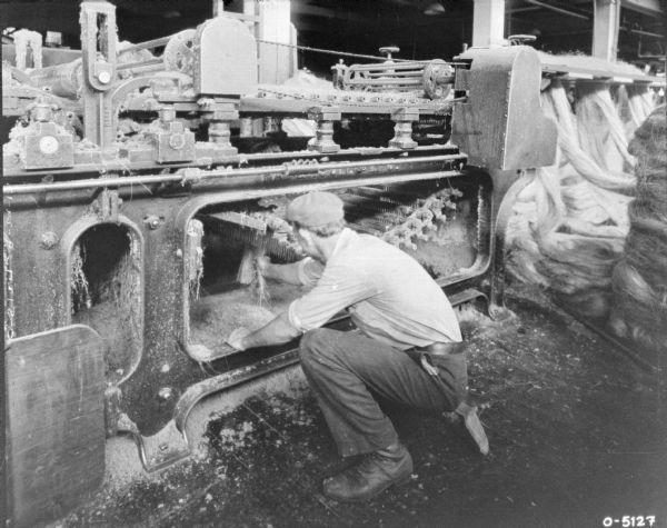 A man is crouching down a holding onto some twine (hemp) in part of the machinery underneath a manufacturing machine indoors on factory floor. 