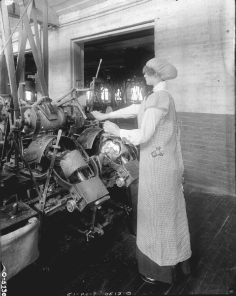 A woman is operating a large, complicated piece of twine machinery. She is wearing a dark skirt and blouse covered by a smock, and her hair is in a large puffy hat. The woman is likely an employee of the International Harvester Osbourne Works in Auburn, New York.