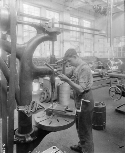 A young man is operating a large mechanical drill. He is dressed in overalls and a work shirt and appears to be rather young. Behind him are several other pieces of machinery. The worker is likely an employee of the International Harvester Osbourne Works in Auburn, New York.