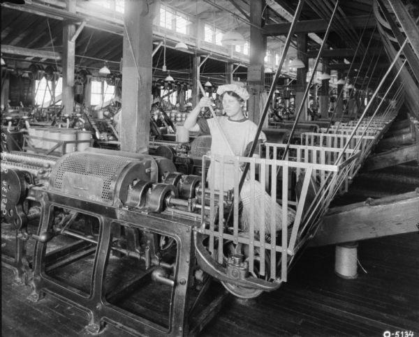 A woman with a broom is standing behind a large piece of twine machinery and is cleaning it with a broom. The woman is dressed in a smock and hat. Behind her the room is filled with twine machinery. This factory may be located at the Osbourne Works International Harvester factory in Auburn, New York.