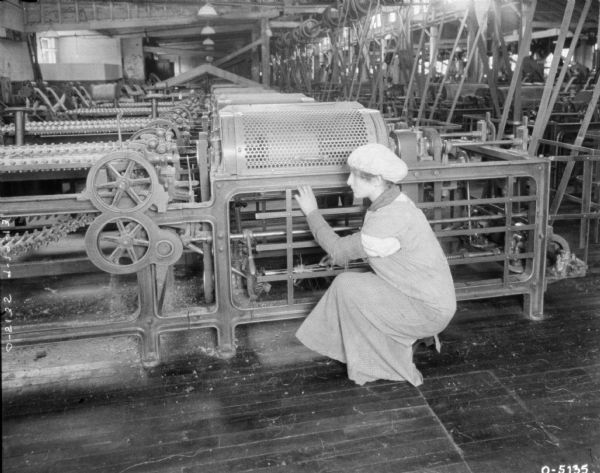 A woman is crouching next to a large piece of twine machinery cleaning it. The woman is dressed in a smock and hat. Behind her the room is filled with machinery. This factory may be located at Osbourne Works in Auburn, New York.