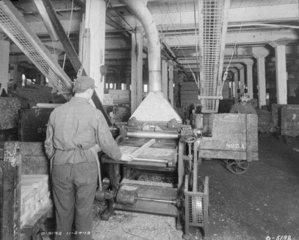 A man is standing with his back to the camera working with a large piece of metal forming machinery. To his right another man is pushing a cart. The men are both dressed in work clothes and hats. In the background are other pieces of machinery and material. The men may be employees of the International Harvester Osbourne Works in Auburn, New York.