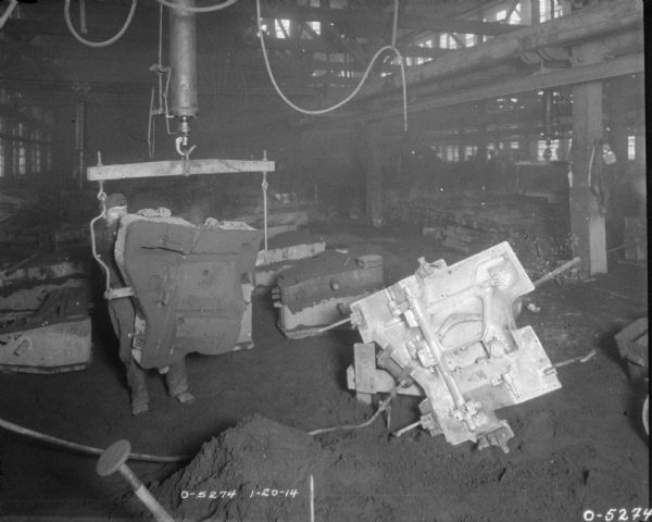 View of a man on factory floor holding onto an engine part that is suspended on a crane. Other engine parts are on the floor in casting forms.