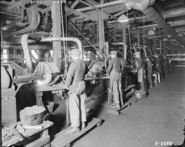 View towards a row of seven men who are standing at metal finishing machinery. The men are wearing work clothes, aprons, and hats, and have their own workstations. Some of the men are standing on raised platforms of wood. The men may be employees of the International Harvester Osbourne Works in Auburn, New York.