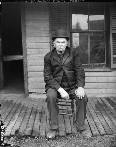 A man, Patrick Gleason, is sitting in a wooden chair on the porch of what appears to be a wood framed house. The man is wearing trousers, a dark shirt, a jacket, and a hat.