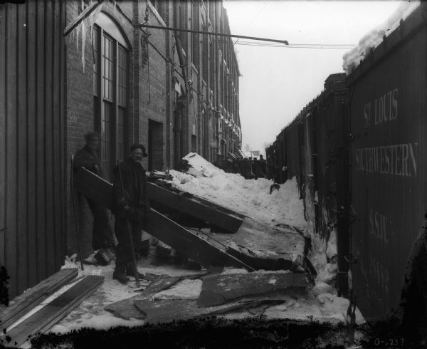 Men are working along the side of a brick building removing snow from against railroad cars.