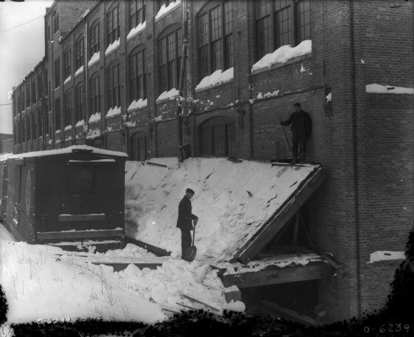Slightly elevated view towards two men working near railroad cars and a brick building. One man is holding a shovel, and the other man is standing at the top of slanted boards pushed up against the building to remove the built-up snow.