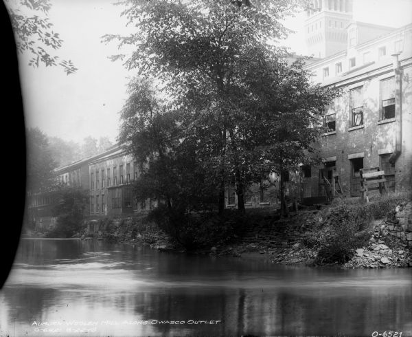 View across water towards mill buildings. Caption at bottom reads: "Auburn Woolen Mill along Owasco outlet."