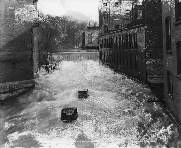 Elevated view looking down at a river and a dam running between brick buildings. Caption reads: "Osborne Works from Genesee Street."