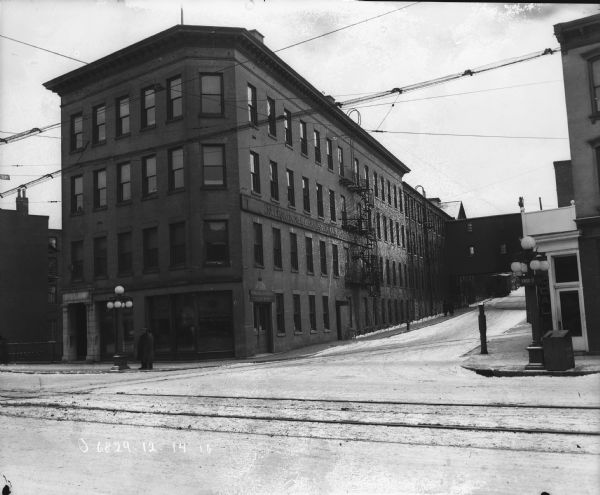 View across Genesee Street towards a brick building and a street or alley. There is a sign on the side that reads: "International Harvester Company of America." A sign in the windows on the front reads: "International Motor Trucks." Snow is on the ground.