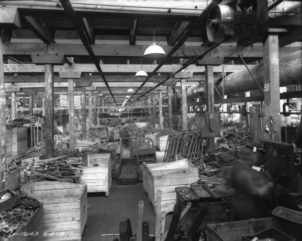 Slightly elevated view of a manufacturing area in a factory. Parts are piled in wood bins, and other parts are stacked on wood tables. A man (blurred by movement) is working in the foreground on the right. An American Flag is hanging from the ceiling in the background.