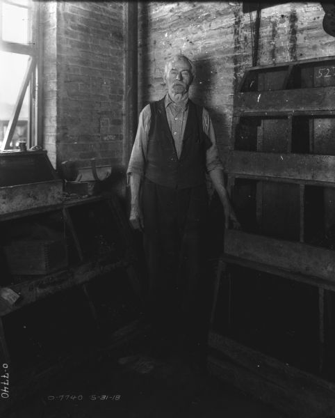 Portrait of an elderly man standing in a corner of a factory near a window with his hand on a bin in a manufacturing area.
