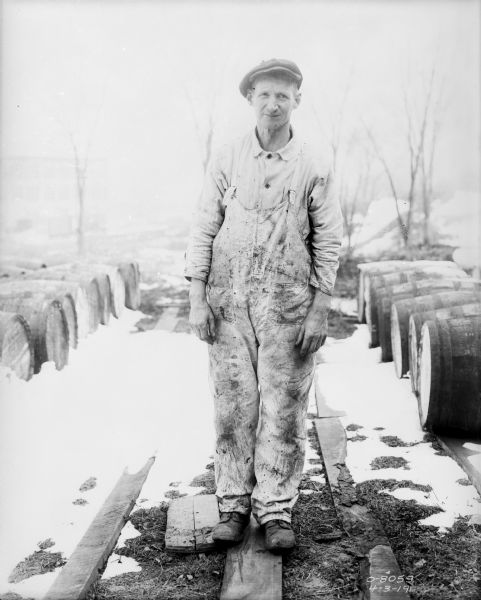 Full-length portrait of a man wearing work overalls and a hat standing outdoors in the snow. Rows of barrels are lying on their sides on wood planks on the left and right.