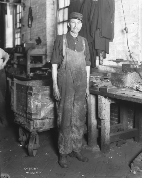 Full-length portrait of a man wearing overalls and a hat. He is standing at a welders bench.
