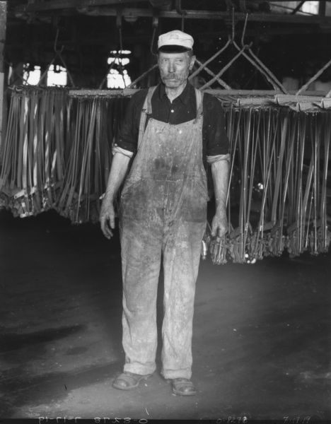Portrait of a man standing indoors wearing overalls and a hat. There are machine parts hanging from hooks behind him. The man is missing parts of his fingers on his left hand.