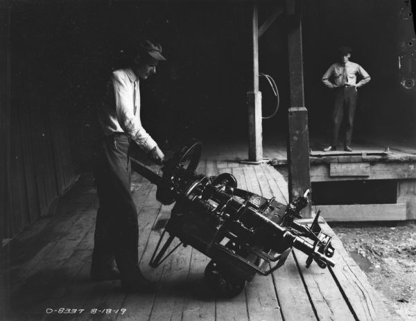 Man moving parts and axles with a delivery cart on what may be a loading dock. Another man is standing in the background on the right.