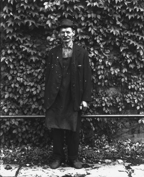 Full-length portrait of a man standing outdoors wearing a hat and eyeglasses, and a business coat over work clothes and an apron. His left thumb is bandaged. The brick wall behind him has leafy vines growing on it.