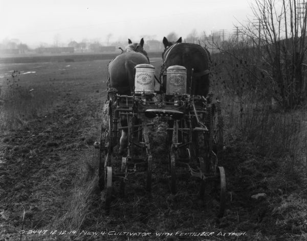 Rear view of a horse-drawn cultivator with fertilizer attachments working in a field. Power lines and a fence are along the field on the right. Buildings are in the distance.
