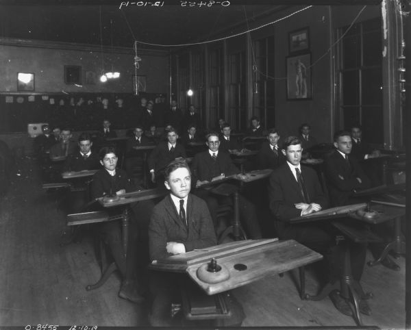 Male students are posing sitting at desks in a classroom. Three men are standing along the back wall near windows.