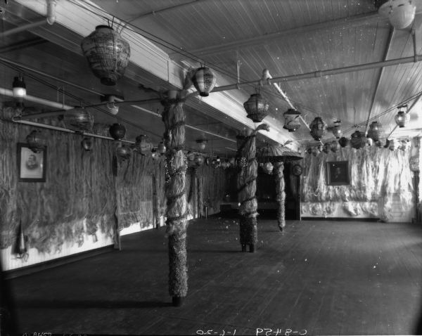 Room decorated for party, probably an employee's New Year's Eve party. Paper lanterns are hanging from the ceiling, and the columns are covered with dried grass or hemp, and dried flowers. There is a portrait of Cyrus Hall McCormick on the wall on the left.
