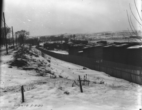 View down side of road towards a lumber yard behind a fence. A road is on the left, with power poles running along side. Railroad cars are on tracks behind the lumber yard, and in the distance are hills.