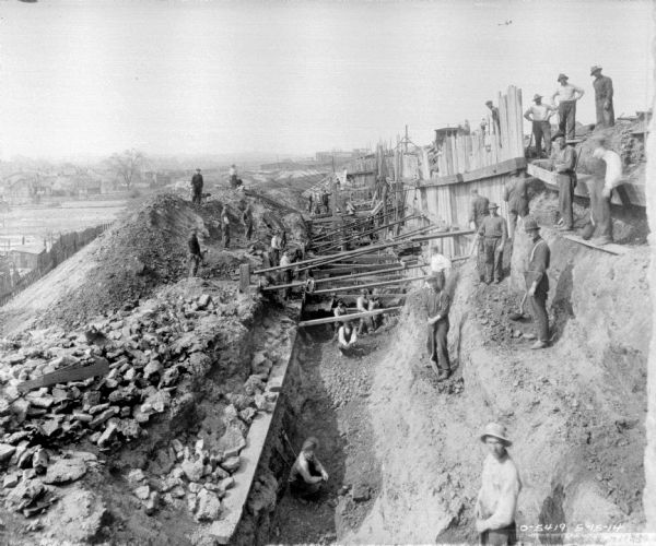 Slightly elevated view of men in and around a trench at a construction site. There is a retaining wall above the trench on the hill on the right. Another fence is further below on the left. Houses are in the distance.
