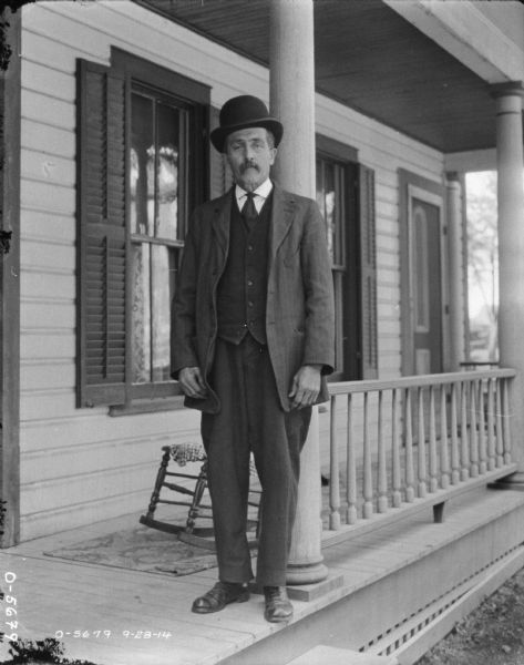 Full-length portrait of Leroy Marsh standing on the porch of a house. He is wearing a suit and hat. 