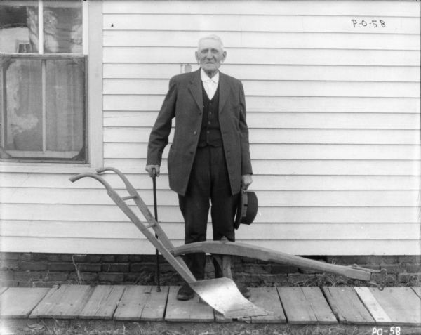Old man with cane in business suit posing with walking plow. He is standing on a wood sidewalk in front of a building. He is holding a hat in his left hand.