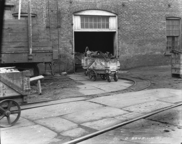 Carts full of materials are near and under an open doorway of a brick building. A railroad car is on tracks on the left.