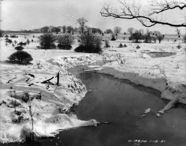 Landscape view of a stream, and harvested cornfield in the distance. Snow is on the ground.
