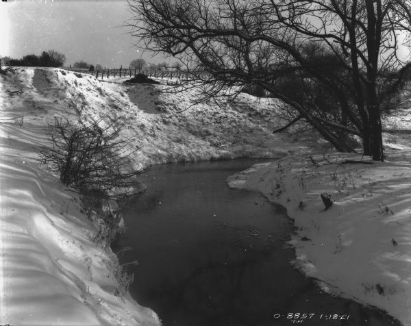 Landscape view of a stream, and harvested cornfield in the distance. Snow is on the ground.